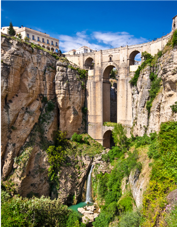 Bridge of Ronda, Spain, spanning a deep gorge with a waterfall, under a blue sky.