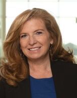 Woman with reddish-blonde hair smiles at the camera; wearing a blue top and black blazer.