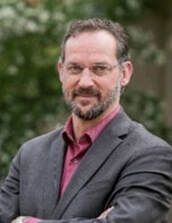 Man with glasses, beard, in a grey suit jacket and red shirt, arms crossed.