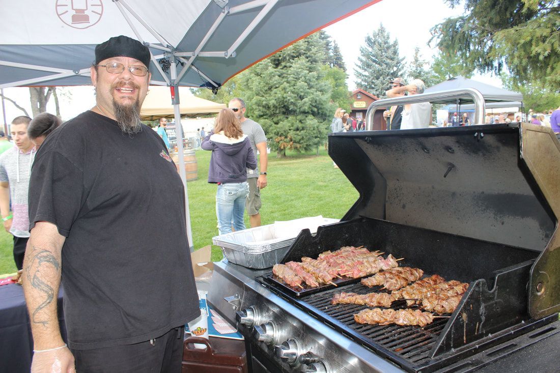 Man grilling meat at an outdoor event. He wears a black shirt and glasses.