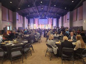 People seated at tables in a large hall, stage in the background, likely a conference or event.