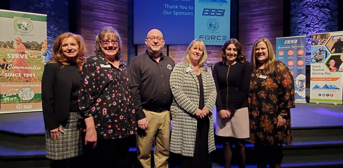Six people standing on a stage, smiling. Two banners are on either side of the group. Dark background.