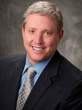 Man with silver hair, smiling, wearing a blue striped shirt, grey tie, and navy suit jacket.