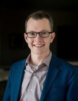 Man with glasses, smiling, wearing a blue blazer, light purple shirt, against a blurred dark background.