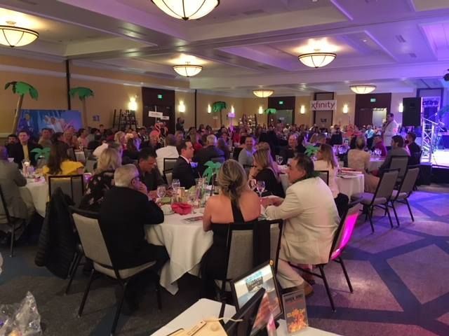 Large event in a banquet hall with many people seated at round tables, band on stage, and tropical decorations.