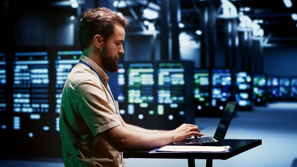 A man is sitting at a desk using a laptop computer in a server room.