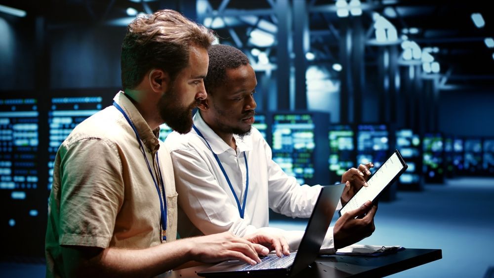 Two men are working on a laptop in a server room.