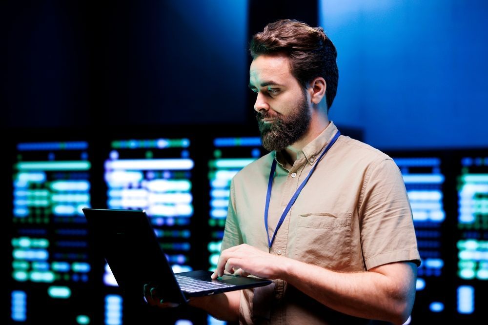 A man with a beard is using a laptop computer in a server room.