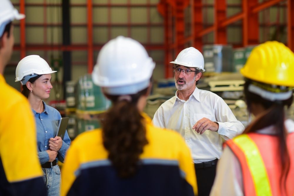 A man is talking to a group of construction workers in a warehouse.