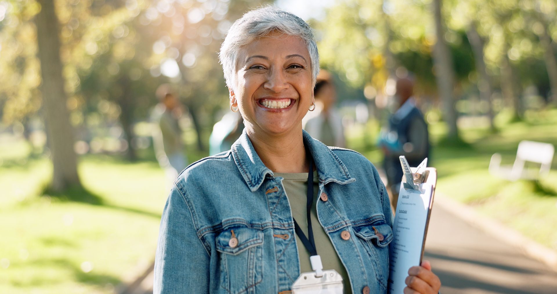 An older woman is smiling while holding a clipboard in a park.