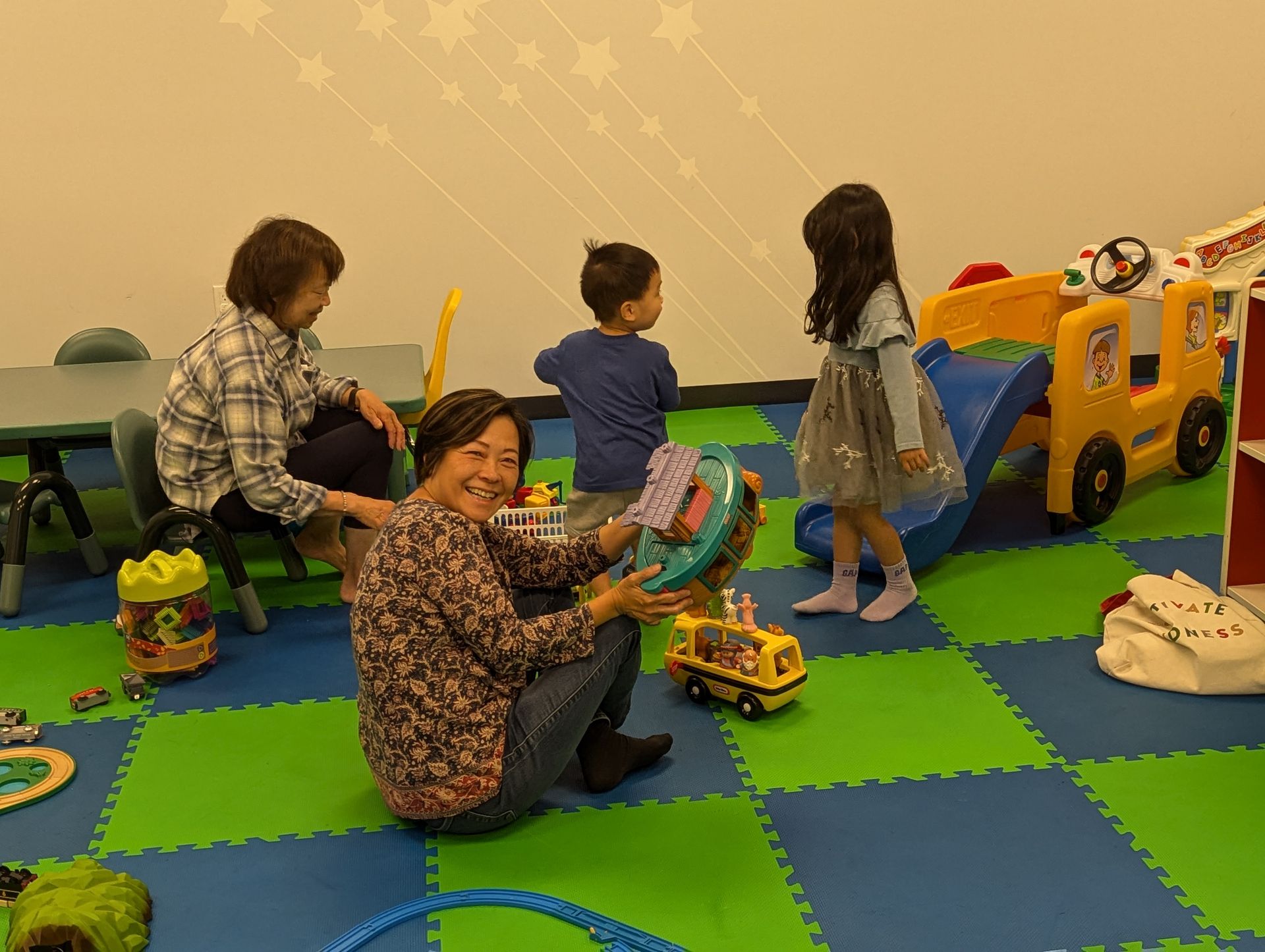 A woman is kneeling on the floor in a play room with children playing with toys.