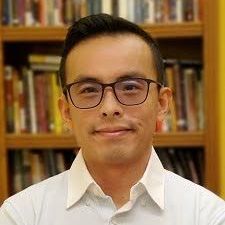 A man wearing glasses and a white shirt is standing in front of a bookshelf.