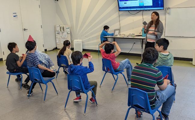 A woman is giving a presentation to a group of children sitting in blue chairs.