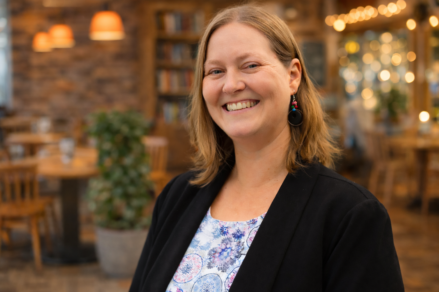 Woman smiles, wearing black blazer over floral top, in a cafe.