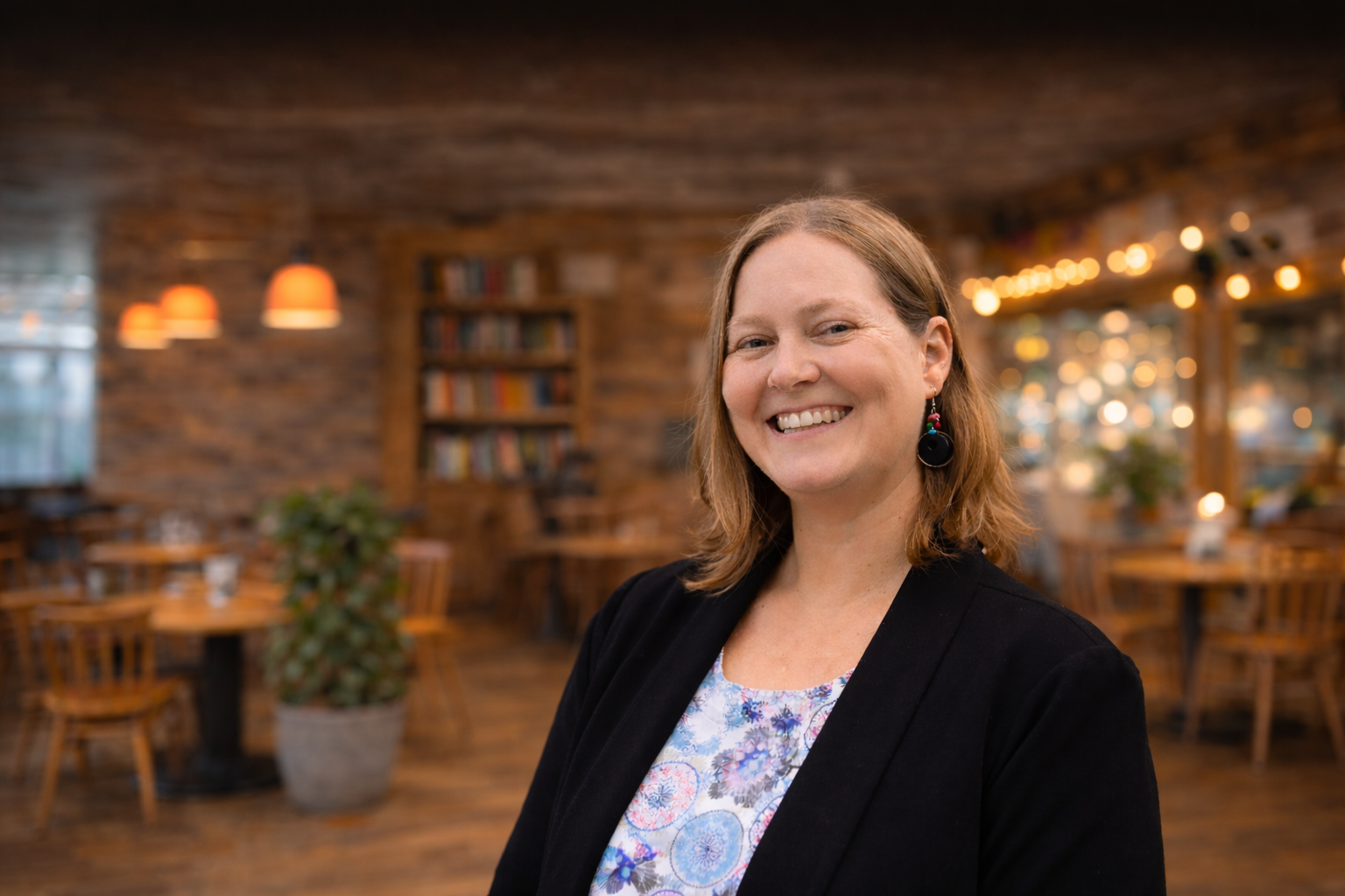 Donna smiling in a cafe, wearing a black jacket over a floral top; brick wall background with tables and lights.
