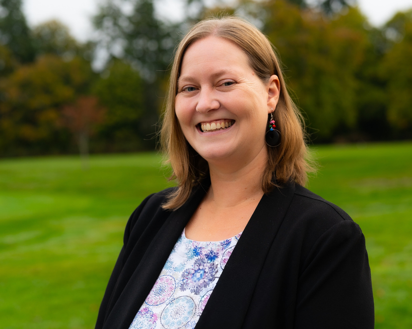 Woman smiling outdoors, wearing black blazer over patterned top, green grassy background.