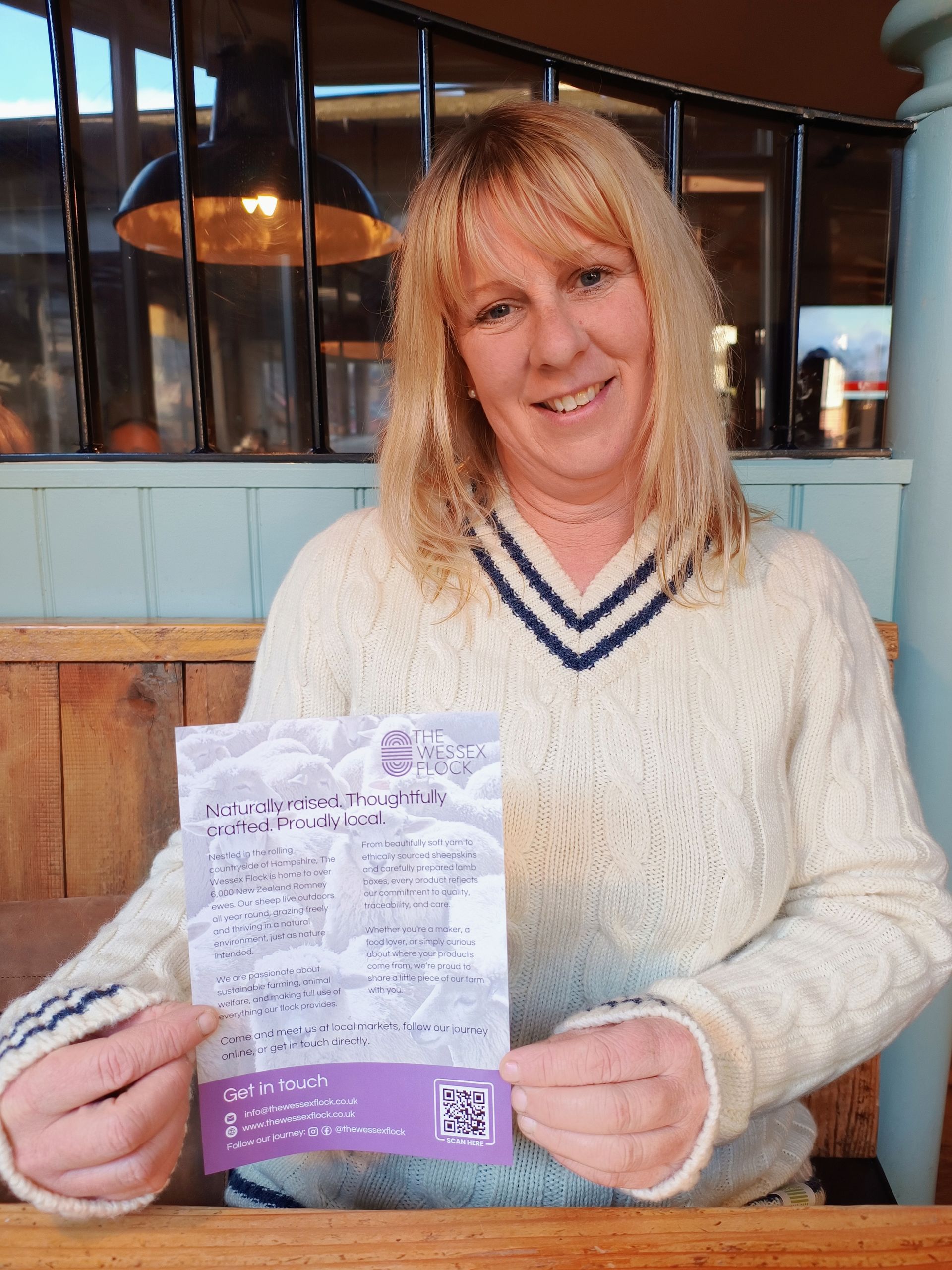A person smiling while holding a printed leaflet featuring a QR code and text, seated at a wooden table in a cafe.