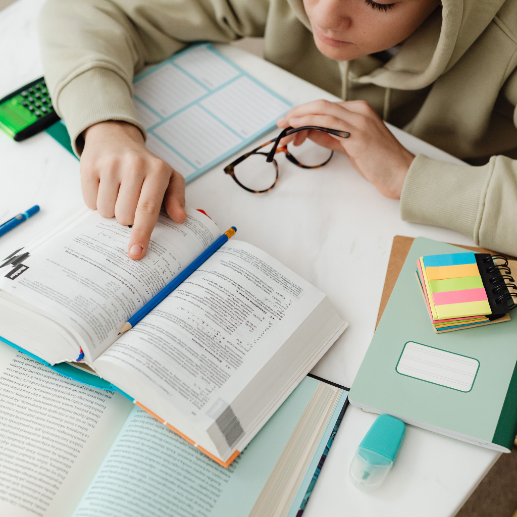 Person studying at a desk with books, notebook, calculator, and glasses.