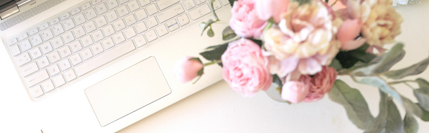 A white laptop keyboard sits next to a soft-focused bouquet of pink and peach flowers on a light-colored surface.