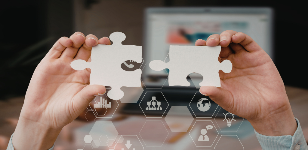 Hands holding two puzzle pieces, with a laptop in the blurred background.