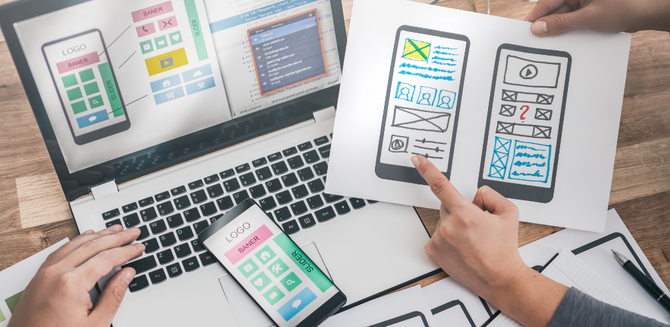 Hands holding tablet, phone, and laptop displaying colorful app wireframes on a desk