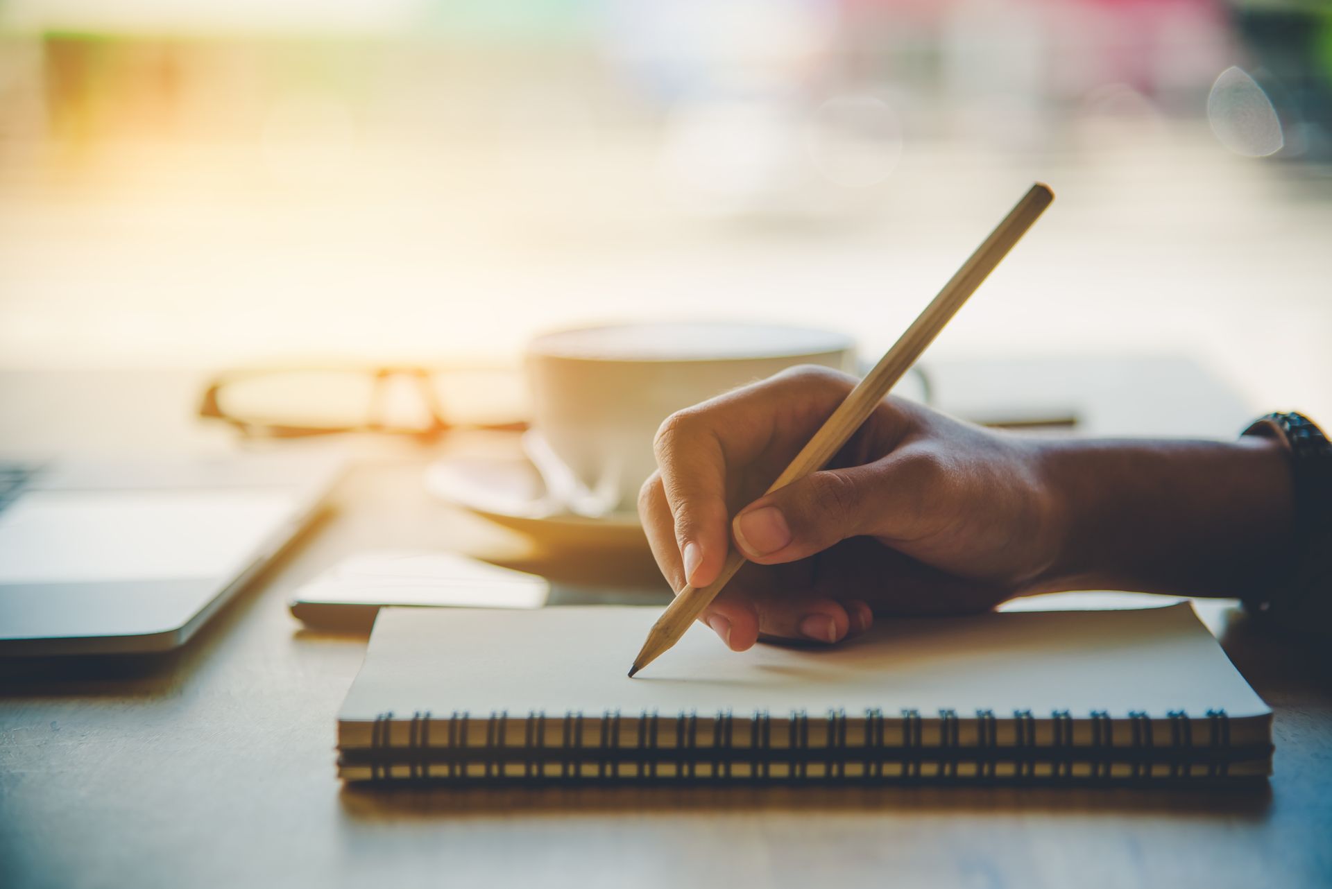 Hand writing in a notebook with a pencil, coffee cup, and glasses on a wooden table.
