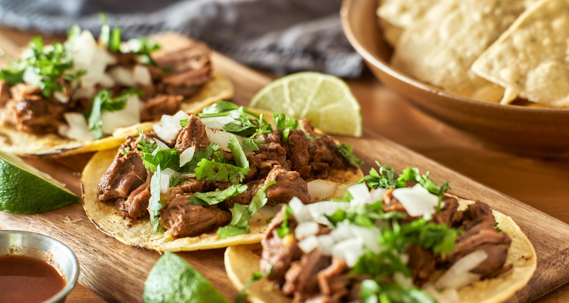 Tacos topped with cilantro, onions, and lime wedges on a wooden board; tortilla chips in a bowl.