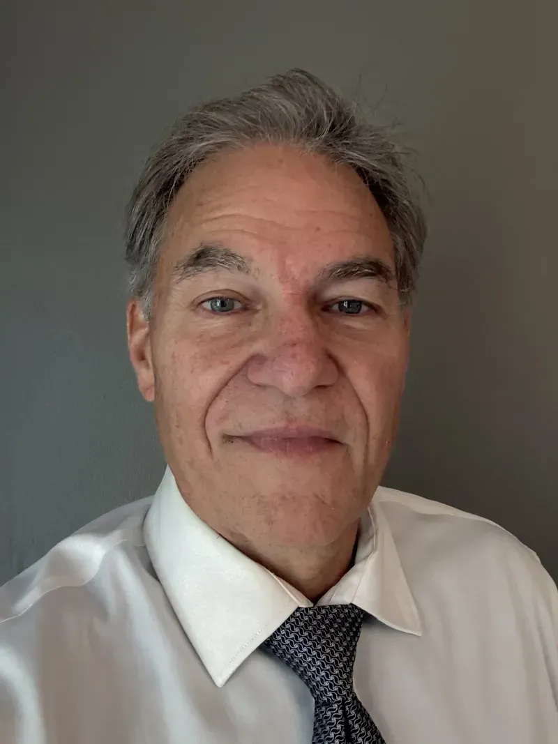Man in white shirt and patterned tie smiles, grey background.