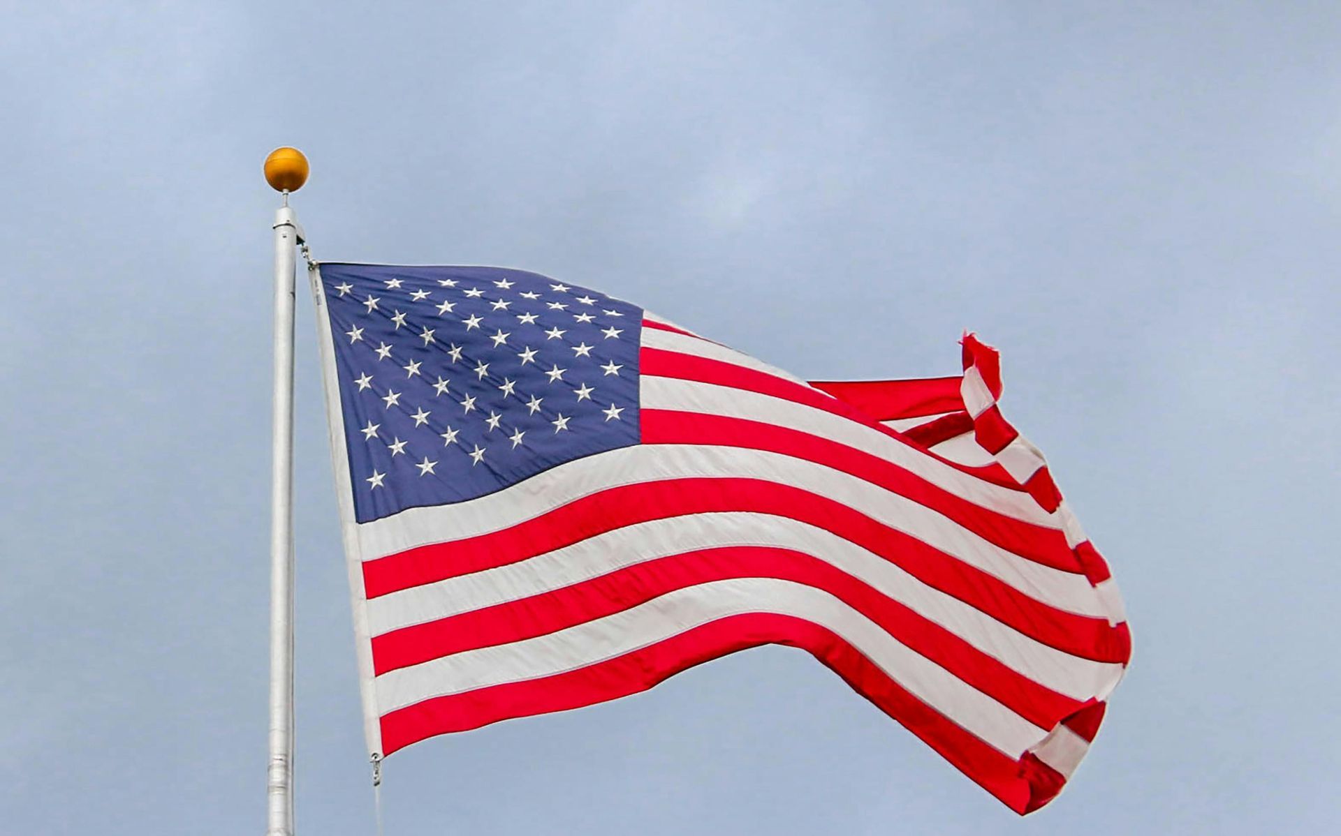 American flag waving in the wind, red and white stripes, blue field with white stars, against a cloudy sky.
