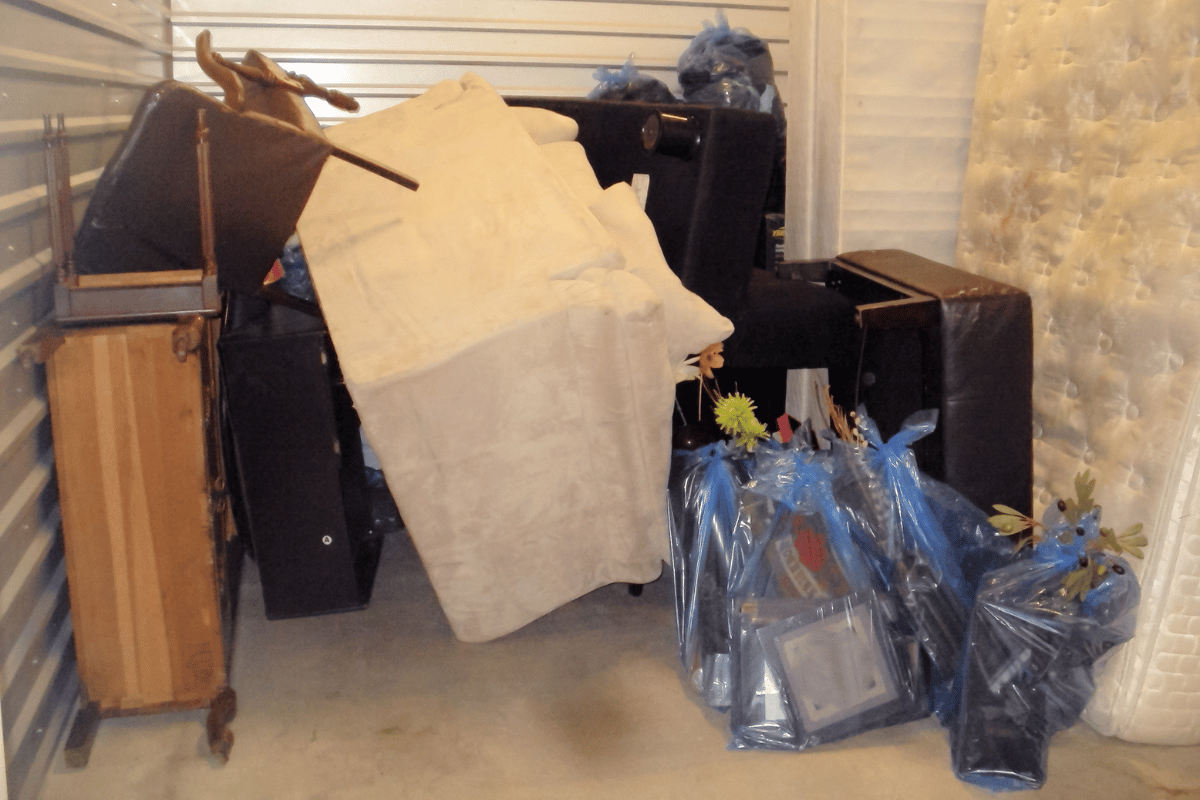 A cluttered storage unit filled with furniture, bags, and a mattress. The contents are a mix of wood, dark, and light-colored items.