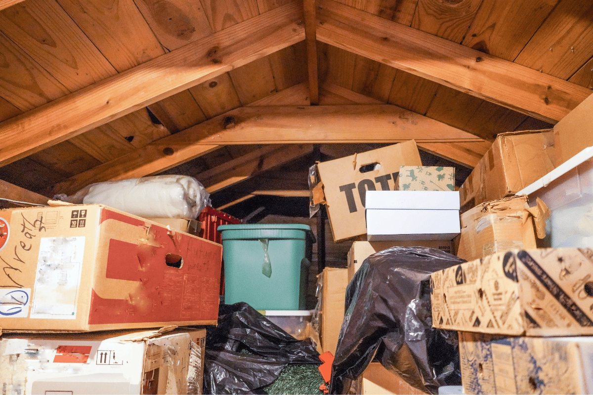 A cluttered attic filled with boxes, bins, and bags under a wooden roof.