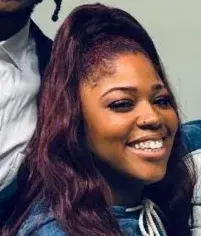 A Black woman with burgundy hair smiles. She wears a blue top and has light skin.