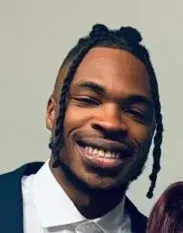 Smiling Black man with dark braided hair, wearing a white shirt and suit jacket.