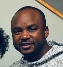 A Black man with a shaved head smiles, wearing a gray hoodie.