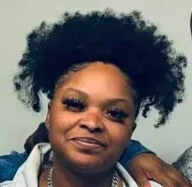 A Black woman with dark, curly hair smiles, wearing a denim jacket and silver chain. She appears to be indoors.