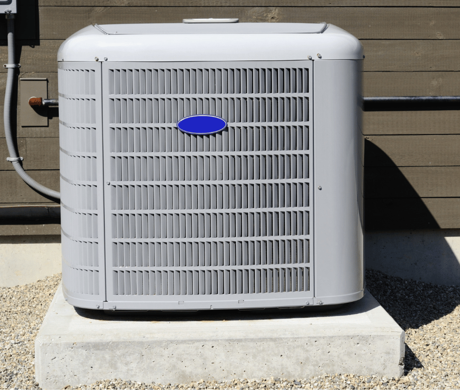 Gray air conditioning unit on a concrete pad, outdoors against a brown wooden wall.
