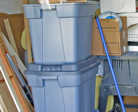 Two blue storage bins stacked in a cluttered garage, with a broom leaning against them.