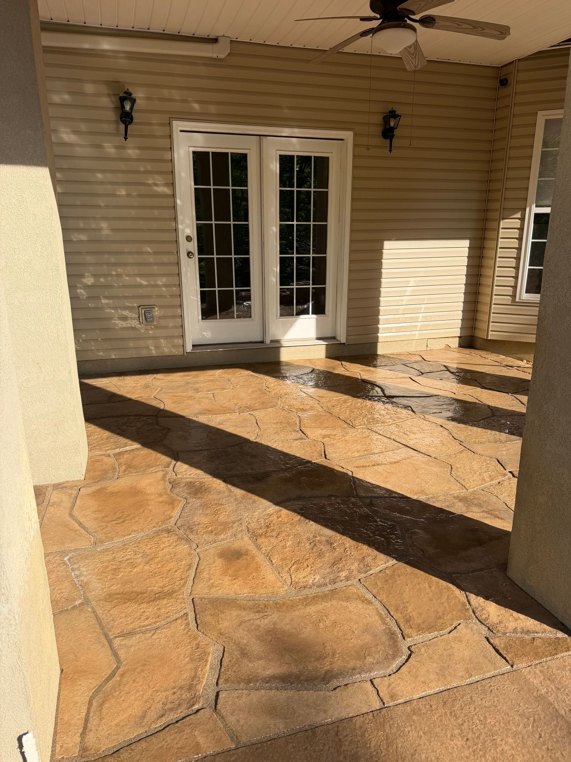 Sunny stone patio with beige siding, white French doors, wall lanterns, and long shadows.