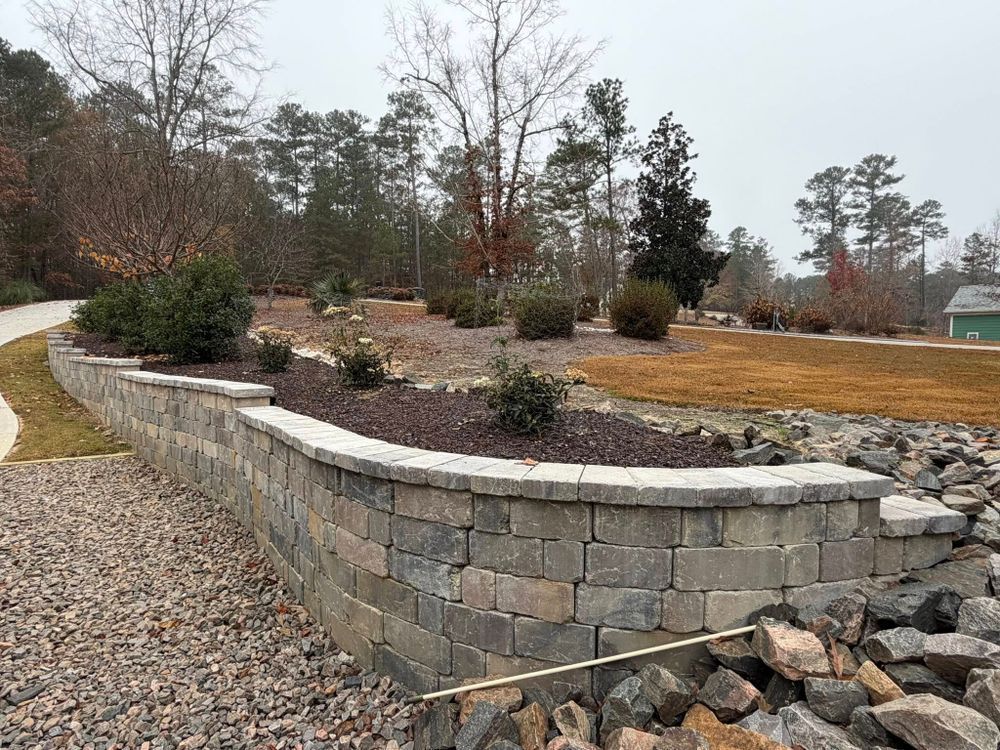 Stone retaining wall with landscaped hillside, shrubs, gravel path, and bare trees under an overcast sky