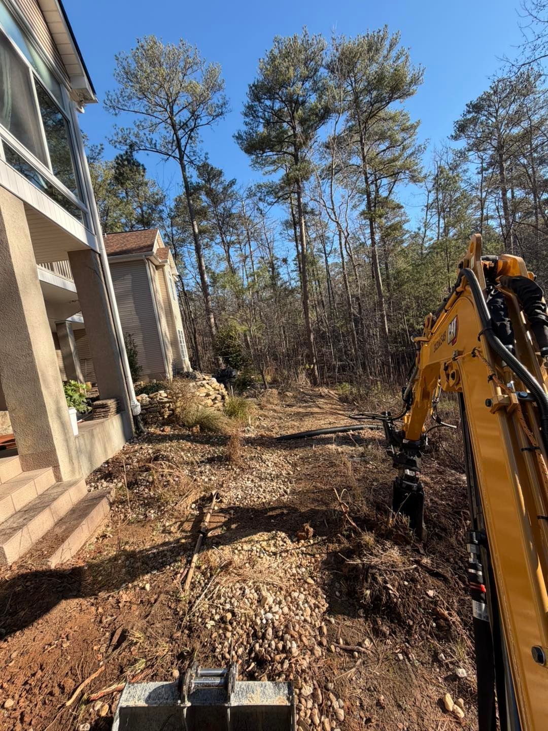 Excavator clearing brush beside a house in a wooded yard under a blue sky