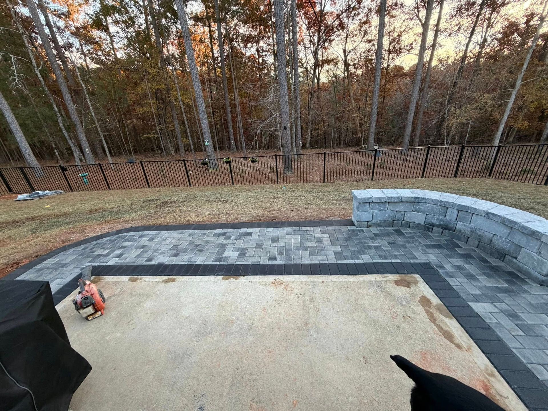 Backyard patio under construction with gray pavers, curved seating wall, and wooded backdrop