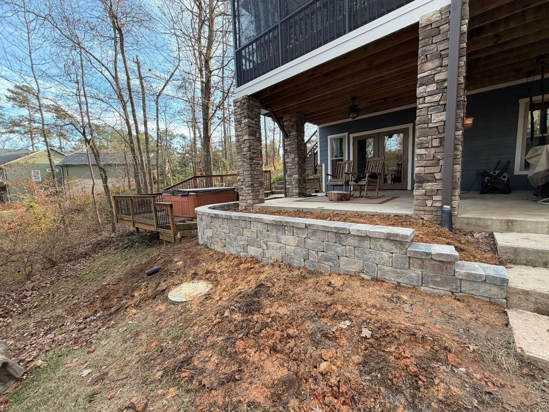 Stone patio and covered porch beside a wooded yard with fallen leaves and bare trees