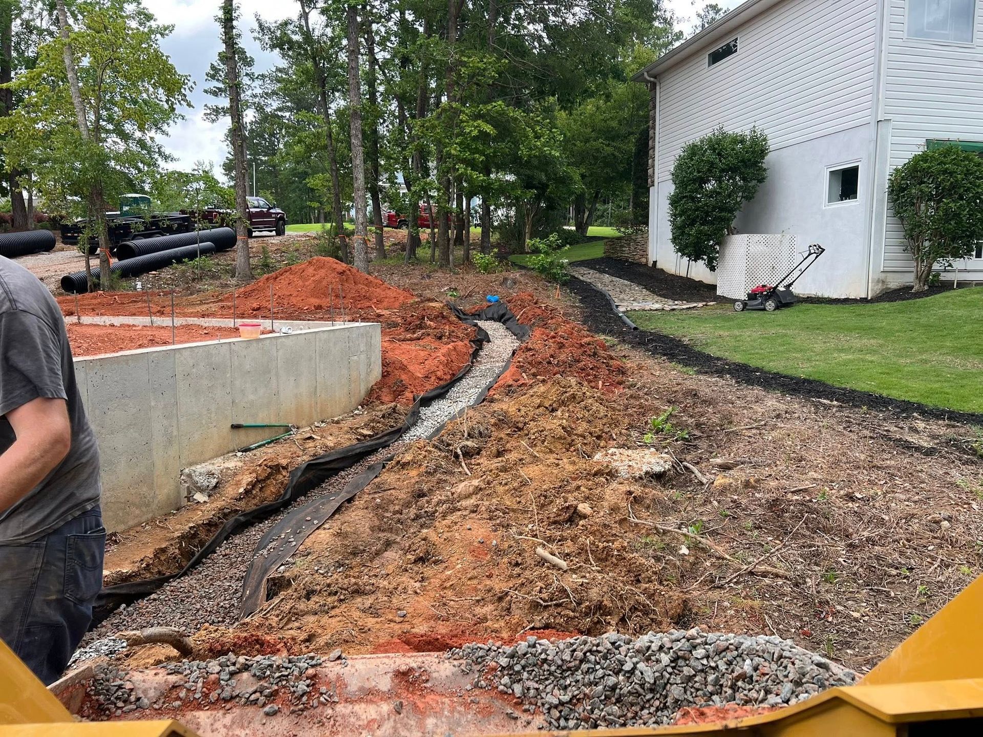 Backyard drainage trench with gravel and orange dirt beside a white house, trees, and a concrete wall