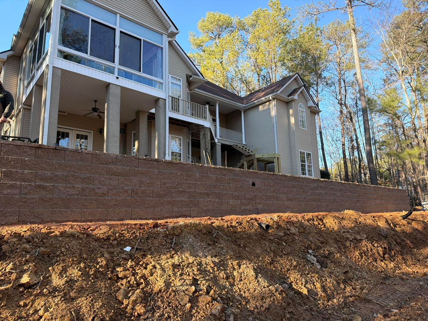 Two-story house with a deck on a sloped dirt lot, bordered by a brick retaining wall and tall trees
