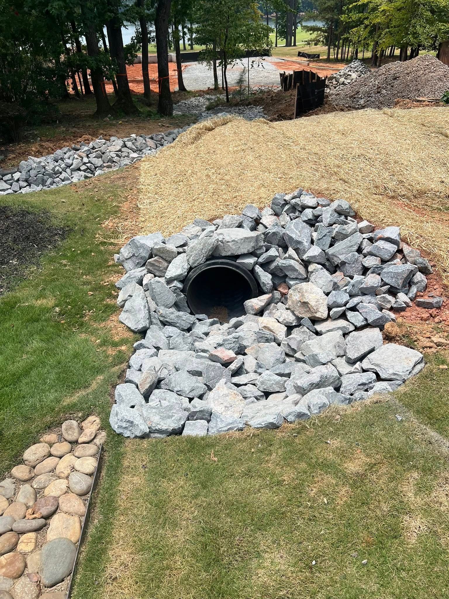 Stone-lined drainage path with gray rocks and gravel in a grassy park area with trees.