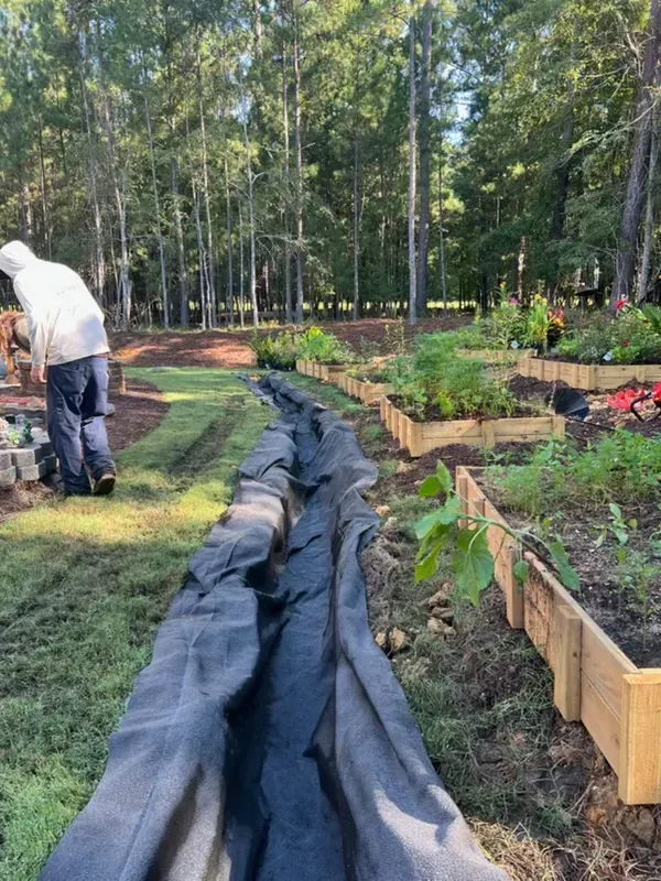 Person working in a garden beside raised beds and a long black landscape fabric row in a wooded area