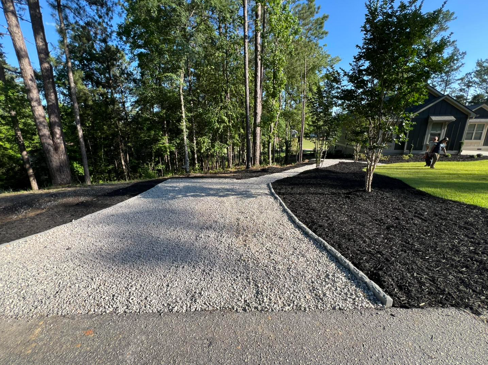 Gravel driveway bordered by mulch and trees leading to a house in a sunny yard