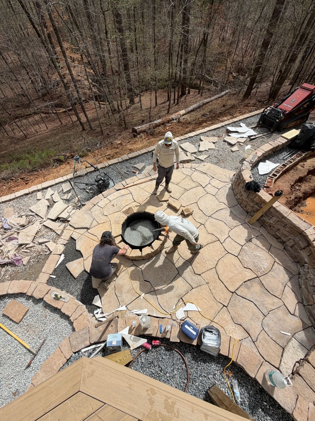 Two workers on a stone patio around a circular fire pit, with forest beyond