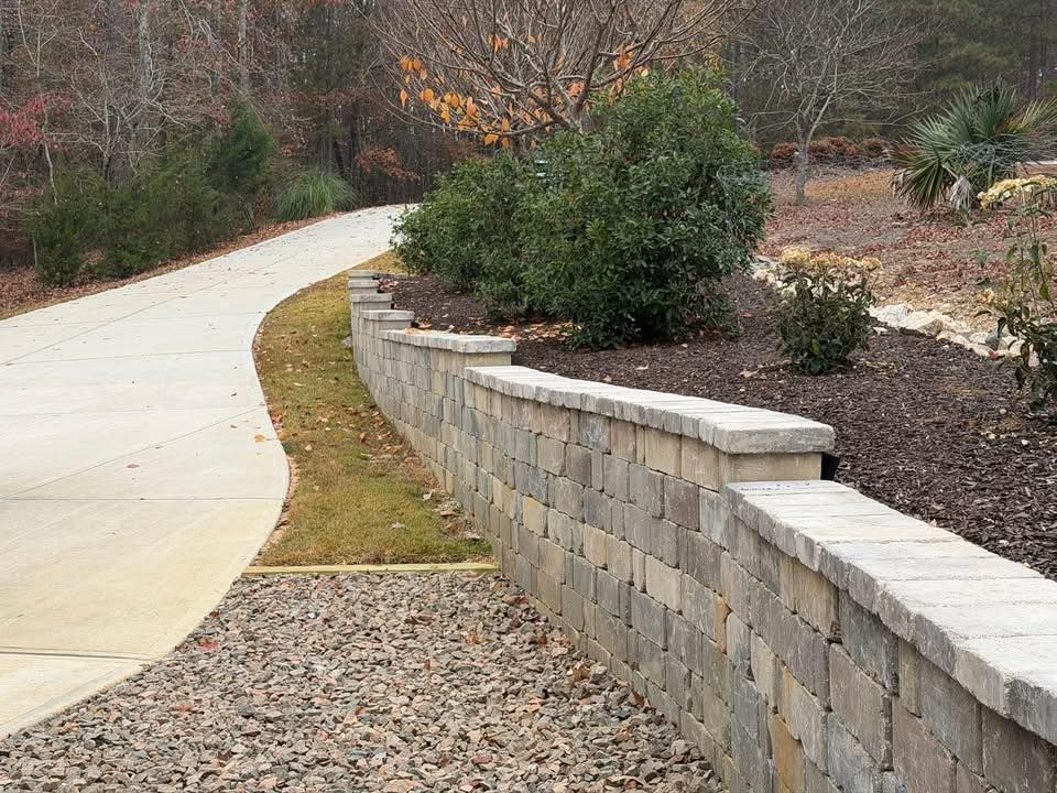 Curved concrete walkway beside a low stone retaining wall and landscaped shrubs on a hillside
