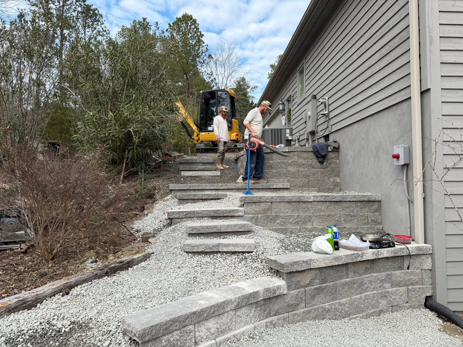 Workers installing steps on a steep concrete hillside beside a gray house, with machinery at the top.